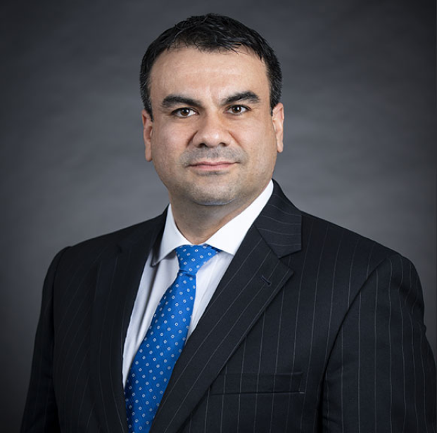 Professional headshot of a man in a dark pinstripe suit with a blue tie, posing against a gray background.