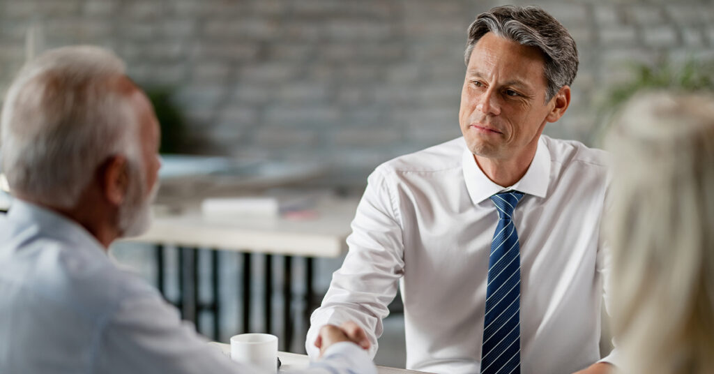 A male professional in a white shirt and tie shakes hands with an older man during a meeting or consultation.