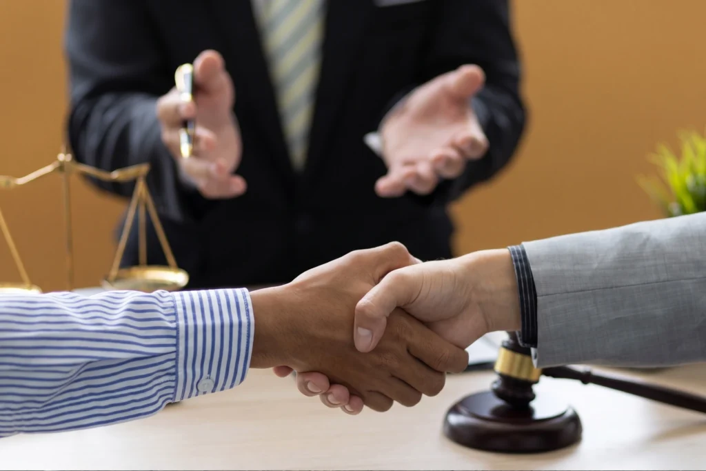 Two people shaking hands in a legal office, with a lawyer, a gavel, and scales of justice in the background.
